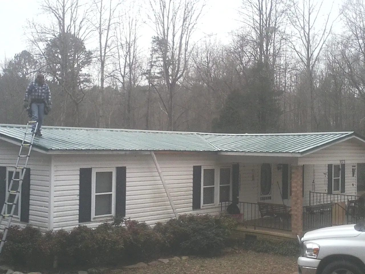 Worker on a ladder beside a new green metal roof on a ranch home in Brockton